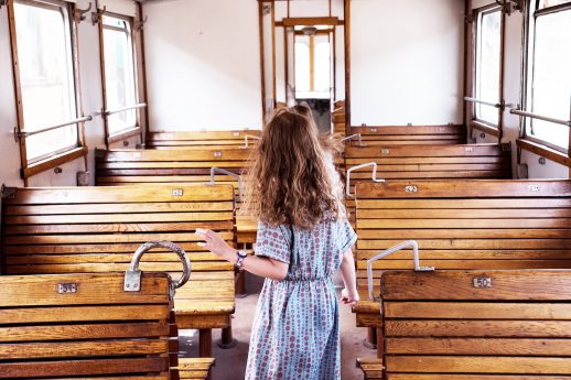 Kobieta w sukience stoi w środku historycznego wagonu kolejowego z drewnianymi ławkami. Jest zwrócona tyłem do kamery, a jej włosy swobodnie opadają na plecy. Wagon jest oświetlony dziennym światłem, które wpada przez okna i podkreśla detal konstrukcji.