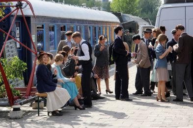Grupa osób ubranych w stylizacje retro gromadzi się na peronie obok pociągu. Mężczyźni i kobiety noszą odzież pasującą do modnych trendów z połowy XX wieku, w tym kapelusze, garnitury i sukienki. W tle widoczne są torowisko oraz kolejne wagony, a scenę otacza przyjemna, słoneczna pogoda.
