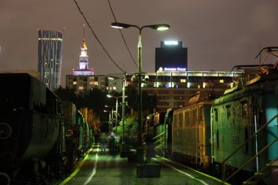 Zdjęcie pokazuje starą stację kolejową nocą, oświetloną ulicznymi latarniami i neonami z pobliskich budynków. Po lewej stronie widać szereg zaparkowanych starych lokomotyw i wagonów kolejowych ustawionych równolegle do siebie. W tle widoczne są nowoczesne wieżowce ze światłami i charakterystycznym zegarem na wieżowcu.