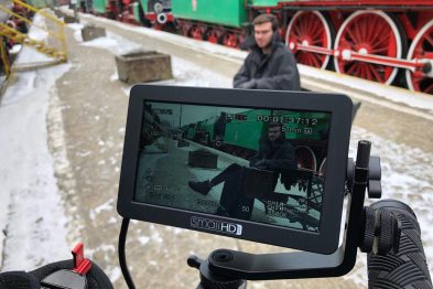 Na pierwszym planie widoczny jest ekran monitora kamery filmowej, który pokazuje mężczyznę siedzącego na schodach zimą. W tle za monitorem widać stojące lokomotywy i wagony kolejowe pokryte śniegiem. Osoba na monitorze jest ubrana w zimowe ubranie, a cała scena ma charakter kolejowy.