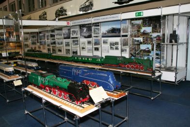 W hali wystawowej prezentowane są modele lokomotyw o różnych kolorach i wzorach, umieszczone na stołach ułożonych w rzędzie. Nad modelami zawieszone są półki z fotografiami i innymi eksponatami kolejowymi. Całość tworzy przejrzystą ekspozycję skierowaną do miłośników kolei i historii transportu.