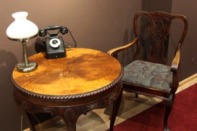 Stolik z politurą i zdobionymi nogami ozdobiony jest lampą z kloszem oraz starym modelem telefonu z krążkiem wybierania numerów. Obok stolika stoi drewniane krzesło z oparciem w kształcie litery 
