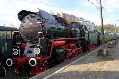 Ogromna, odrestaurowana czarno-czerwona lokomotywa parowa stoi na torach w towarzystwie innych zielonych wagonów kolejowych. Maszyna ma liczne białe akcenty, w tym koła i obwódki, oraz wyraźnie zaznaczoną czarną przednią część z dużą okrągłą lampą. W tle widoczne są drzewa i fragmenty innych pojazdów kolejowych.