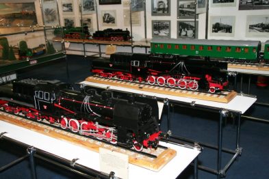 Modele lokomotyw stoją na wystawie obok siebie na platformach, są koloru czarnego i zielonego z czerwonymi elementami. Za modelami widoczne są fotografie i opisy dotyczące historii kolejnictwa. W tle mieszczą się witryny z kolejnymi eksponatami związanymi z tematyką pociągów.