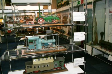 W gablotach eksponowane są modele pociągów i elementy związane z kolejnictwem. Obok modeli widoczne są opisy w języku polskim. W tle dostrzec można inne eksponaty oraz odwiedzających muzeum.