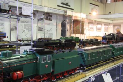 Widoczne są modele lokomotyw kolejowych umieszczone na makiecie torów. W tle za szklanymi witrynami znajdują się eksponaty i informacyjne tablice. Całość prezentuje się estetycznie i jest dobrze oświetlona, co sugeruje profesjonalne podejście do ekspozycji.