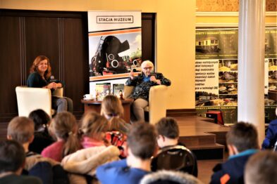 Dwoje dorosłych siedzi na fotelach w pomieszczeniu, które wygląda na salę konferencyjną lub czytelnię. Osoba z mikrofonem w dłoni, ubrana w szalik i białe okulary, wydaje się opowiadać historię dzieciom siedzącym na podłodze przed nią. W tle widoczne są modele pociągów i kolejowa tematyka na plakacie.
