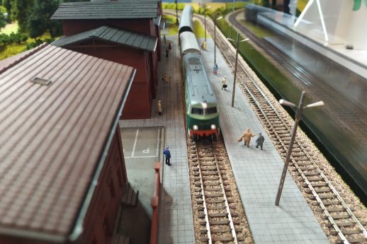 Makieta kolejowa ukazuje miniaturową scenę z pociągiem, torami i peronem; widać także modele ludzi i budynków. Kolorowy pociąg zlokalizowany jest na środku kompozycji, flankowany przez dwa wiernie odwzorowane, czerwone budynki stacji kolejowych. Sceneria obejmuje elementy krajobrazu, takie jak drzewa i krzewy, co nadaje makiety realistyczny charakter.