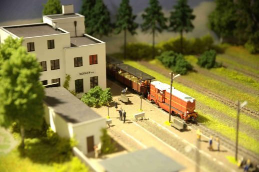 Model pociągu w barwach czerwono-białych stoi na miniaturowym peronie obok dwupiętrowego budynku, przypominającego stację kolejową. Dookoła rozciąga się zielony krajobraz z modelami drzew, a tory kolejowe przebiegają przez środek kompozycji. Postacie ludzkie są rozmieszczone wokół peronu i budynku, dodając realizmu do sceny.