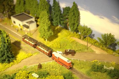 Model pociągu retro przemierza malowniczy krajobraz na makiecie kolejowej, mijając drogę, drzewa i budynki. Makieta odwzorowuje wiejski teren z detalami takimi jak pojazdy, ogródek i figurki ludzi. Ciepła kolorystyka oraz staranne wykonanie elementów makiety podkreślają urok miniatury krajobrazu kolejowego.