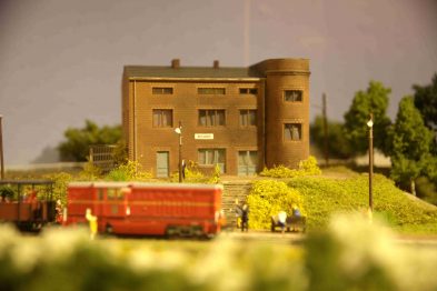 Makieta kolejowa przedstawia model budynku stacji z charakterystyczną wieżą wodną obok. Po lewej stronie widać model pociągu towarowego z czerwoną lokomotywą, który stoi na torach. Makieta zawiera roślinność oraz drogę, po której porusza się żółty pojazd, dodając realizmu scenie.