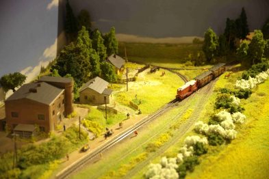 Makieta kolejowa przedstawia krajobraz wiejski z domami, roślinnością oraz pociągiem na torach. Na ścieżkach widać detale takie jak modele ludzi i pojazdów. Oświetlenie modelu tworzy efekt naturalnego światła słonecznego.