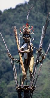 Osoba ubrana w tradycyjne szaty stoi na wysokiej drewnianej konstrukcji. Na głowie ma ozdobny hełm z piórami, a w ręce trzyma długie, cienkie przedmioty. W tle widać zielone wzgórza porośnięte gęstą roślinnością.