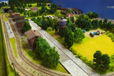 Model kolejowy przedstawia pejzaż z torami kolejowymi, mostem i budynkami. Widać także drzewa, które otaczają torowisko i mały budynek stacyjny. Model zawiera też elementy krajobrazu jak pola i łąki oraz wodę symulującą jezioro lub rzekę.