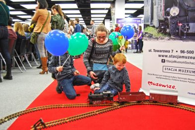 Dwoje dzieci wraz z dorosłym siedzi na czerwonym dywanie, obok modelu torowiska z miniaturowym pociągiem. Jedno z dzieci trzyma niebieski balon z napisem 
