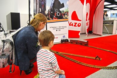 Kobieta klęczy obok małego chłopca na czerwonym dywanie, obserwując model kolejki, który porusza się po torach na miniaturowym makiecie. Za nimi znajduje się namiot z logo województwa mazowieckiego oraz baner informacyjny o targach. W tle widoczne są elementy wystawiennicze i inni odwiedzający targi.