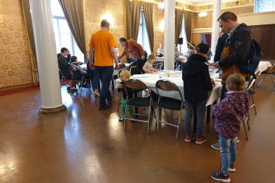 Grupa osób zgromadzona jest wokół stołów, na których rozłożone są kartonowe modele i narzędzia do sklejania. Dorośli i dzieci są skupieni na pracy, wydają się być zaangażowani w składanie modeli. Pomieszczenie, w którym się znajdują, jest jasne z dużymi oknami i klasyczną dekoracją wnętrz.