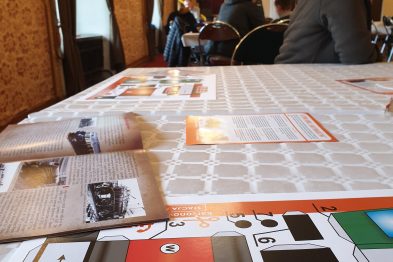 Na długim stole rozłożone są kartonowe elementy modeli pociągów. W tle widać skupione osoby siedzące przy podobnych stołach z kartonowymi modelami. Elementy mają wydrukowane szczegóły takie jak okna czy koła, w gotowości do wycięcia i sklejenia.