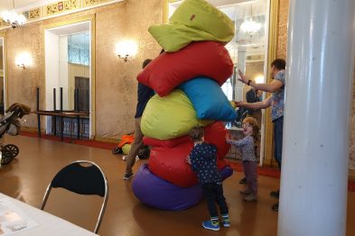 W przestronnym pomieszczeniu z widocznymi zdobieniami na suficie dzieci bawią się kolorowymi, dużymi poduszkami ułożonymi w stos. Osoby dorosłe znajdują się w tle, jedna z nich trzyma w ręku poduszkę, a inna stoi obok. Na pierwszym planie widoczne jest małe dziecko w kolorowych ubraniach, stojące obok stosu poduszek oraz odwrót krzesła.