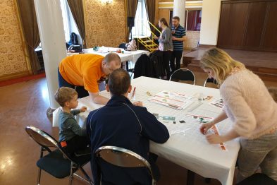 Osoby siedzą przy białych stołach i składają modele z kartonu; widoczne są kartki, nożyczki i kleje. W tle stoją kolejne osoby oraz eksponaty kolejowe. Uczestnicy zajęciów koncentrują się na swoich projektach, pracując indywidualnie lub wspólnie.