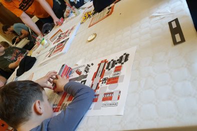 Osoby siedzą przy dużym stole i składają modele z kartonu, koncentrując się na swoich projektach. Na stole rozłożone są arkusze z wydrukowanymi częściami modeli pociągów do wycinania i sklejania. Dziecko w centrum zdjęcia trzyma w rękach częściowo złożony model czerwonego wagonu.