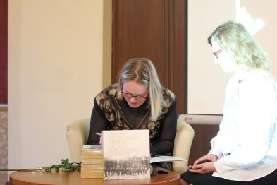 Dwie osoby siedzą przy stole w pomieszczeniu o klasycznym wykończeniu, jedna z nich w trakcie czytania lub podpisywania książki, druga obserwuje ten proces. Na stole leży stos książek, a wśród nich jedna otwarta, przed nią znajduje się długopis. Światło zewnętrzne wpada do środka przez duże okna, tworząc przyjemną atmosferę.