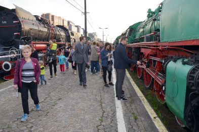 Zdjęcie przedstawia grupę ludzi zwiedzających wystawę związaną z kolejnictwem, zatrzymując się przy eksponowanych lokomotywach do oglądania ich z bliska. Wśród zwiedzających dominują dorośli, ale widoczne są również dzieci. Eksponowane lokomotywy są w różnych kolorach, a na pierwszym planie widać zieloną maszynę z czerwonymi elementami.