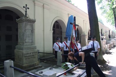 Grupa osób uczestniczy w ceremonii składając kwiaty na grobie. Niektórzy trzymają flagę Polski i sztandar z emblematem kolejowym. Są ubrani w białe koszule i czarne spodnie, a otoczenie to zacieniony teren cmentarny.