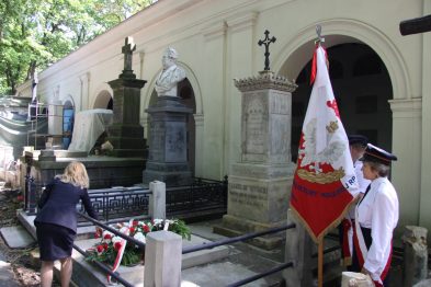 Grupa osób stoi przy grobowcu ozdobionym wieńcami i kwiatami. Osoba w białej koszuli i czarnej czapce trzyma flagę z herbem. Grobowiec przykryty jest ciemnym dachem, a w tle widoczne są drzewa i nagrobki.