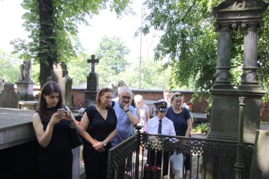 Grupa osób stoi otoczona drzewami i nagrobkami na cmentarzu, skupiając się wokół jednego grobu. Wśród zebranych widać mężczyznę trzymającego mikrofon oraz kobietę robiącą zdjęcie smartfonem. W tle dostrzec można żeliwną balustradę oraz pomnik nagrobny.