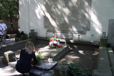 Grupa osób skupia się wokół grobu z kwiatami na przykładem dwóch klęczących osób układających bukiety. Mężczyzna w jasnej koszuli stoi z lewej strony, obserwując scenę. Cień drzew tworzy na ścianie nagrobka i otaczających go ławek kontrast w stosunku do oświetlonej słońcem sceny.