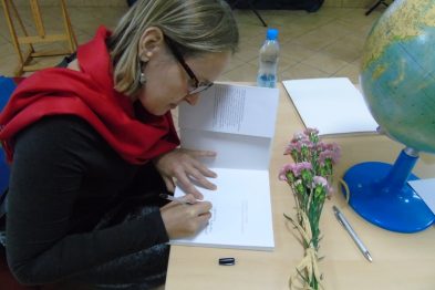 Kobieta w czerwonym szaliku siedzi przy stole i coś pisze. Przed nią na stole leży otwarta książka, obok niej stoi butelka wody i leży długopis. W tle widać globus oraz kwiaty na biurku.