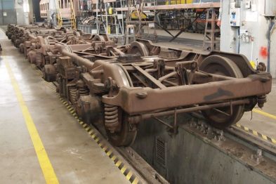 Szereg wózków kolejowych ustawiony jeden za drugim w hali naprawczej. Każdy wózek składa się z dwóch par kół i metalowej konstrukcji połączeniowej. Na posadzce znajdują się oznaczenia robocze, a tło stanowią narzędzia i maszyny warsztatowe.