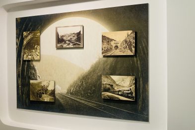 Na ścianie powieszony jest zestaw starych fotografii przedstawiających tematykę kolejową; są one umieszczone w nieregularnym układzie na większym, owalnym tle, które ma dodatkowo dwie prostopadłe linie nawiązujące do torów kolejowych. Każda fotografia jest w ramce, a całość jest oświetlona zapewniając dobry widok na prezentowane treści. Scenę uzupełnia metalowy, biały kształt ramy przypominający wagon kolejowy, w którym ekspon