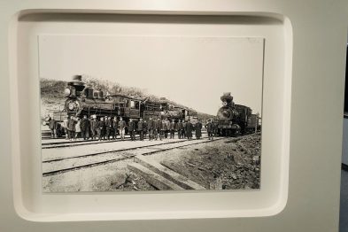 Stara czarno-biała fotografia przedstawia grupę osób stojących obok parowej lokomotywy i zestawu wagonów na torach. Osoby są ubrane w odzież z epoki, wskazującą na koniec XIX lub początek XX wieku; mężczyźni noszą kapelusze i garnitury. Lokomotywa i wagony są szczegółowo widoczne, stoją na prostym odcinku torów.