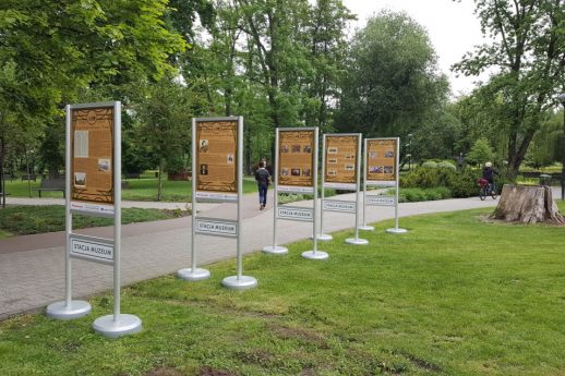 Sześć prostokątnych paneli ekspozycyjnych stoi w rzędzie na trawniku w parku. Panele są umocowane na metalowych stojakach i zawierają informacje oraz ilustracje. W tle widoczne są drzewa i ścieżka parkowa, a na jednym z paneli widnieje tekst.