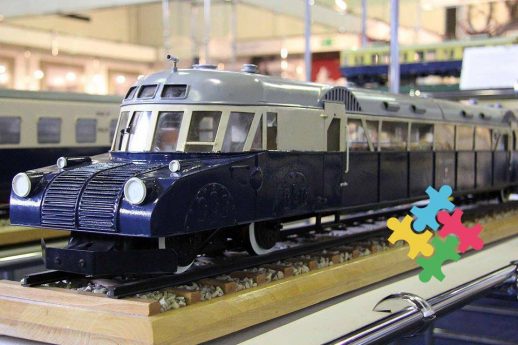 Model ciemnego koloru lokomotywy spalinowej stoi na makiecie torów, otoczony przez inne modele kolejowe. Na pierwszym planie widoczne są trzy kolorowe puzzle sensoryczne ułożone na makiecie. Tło stanowi jasne wnętrze, w którym umieszczone są kolejne elementy modelarskie i eksponaty kolejnictwa.