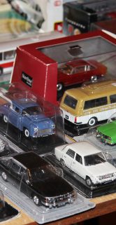 Widoczne są szczegółowe modele samochodów rozmieszczone na stole, niektóre z nich znajdują się w przezroczystych opakowaniach. Modelowe pojazdy mają różne kolory i rodzaje, w tym osobowe, dostawcze oraz ciężarowe. Stoisko z modelami usytuowane jest wewnątrz pomieszczenia, co wskazuje na miejsce spotkania kolekcjonerów i miłośników modelarstwa.