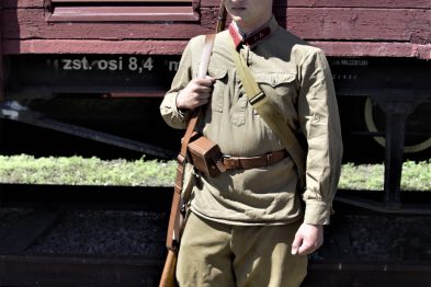 Osoba w mundurze stoi przed towarowym wagonem kolejowym. Na wagonie widnieje biała tabliczka z czarnym tekstem. Osoba trzyma w ręku drewniany karabin i nosi czapkę z daszkiem.