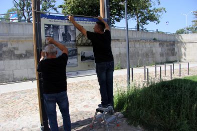 Dwóch mężczyzn montuje wystawę na zewnątrz; jeden stoi na niewielkiej drabinie, a drugi podtrzymuje tablicę z fotografiami. W tle znajduje się kamienny mur, a obok nich widać zieloną trawę. Instalowana tablica zawiera czarno-białe zdjęcia i tekstu nawiązujące do tematyki mostów kolejowych.