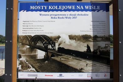 Stara fotografia przedstawia parowóz przekraczający stalowy most kolejowy. Po bokach mostu widoczne są konstrukcje wsporcze. Plakat z informacjami o wystawie umieszczony jest na metalowym stelażu.