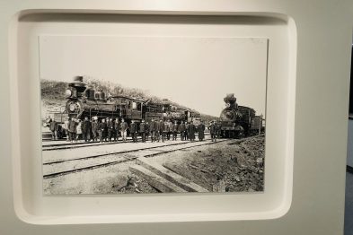 Czarno-biała fotografia przedstawia stary parowóz na torach kolejowych z grupą ludzi w epokowym ubiorze zgromadzonych wokół. Lokomotywa i osoby są ustawione równolegle do torów, na których stoi pojazd. Ekspozycja jest częścią wystawy poświęconej historii kolei w kontekście miasta Harbin i Kolei Wschodniochińskiej.