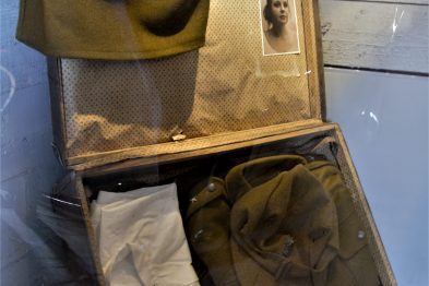Stara walizka stoi otwarta, a w jej wnętrzu znajdują się różne przedmioty, w tym dokumenty i ubranie. Na wewnętrznej stronie pokrywy walizki widać czarno-białą fotografię osoby. Walizka jest częścią ekspozycji, która może oznaczać uwiecznienie wspomnień lub historii związanych z okresem II wojny światowej.