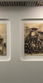 Na ścianie zawieszone są dwa oprawione czarno-białe zdjęcia. Zdjęcie po lewej stronie ukazuje budynek stacji kolejowej, po prawej grupę ludzi na tle drzew i budynku. Oświetlenie galerii rzuca równomierny blask na obie fotografie oraz ścianę.