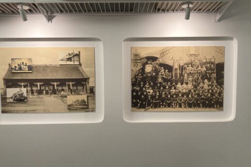 Na ścianie zawieszone są dwa oprawione czarno-białe zdjęcia. Zdjęcie po lewej stronie ukazuje budynek stacji kolejowej, po prawej grupę ludzi na tle drzew i budynku. Oświetlenie galerii rzuca równomierny blask na obie fotografie oraz ścianę.