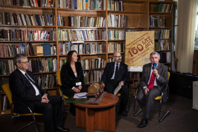 Cztery osoby siedzą przed mikrofonami w pomieszczeniu wypełnionym książkami; dwóch mężczyzn po lewej i kobieta z mężczyzną po prawej. Mężczyzna po prawej trzyma mikrofon i coś mówi, pozostali uczestnicy są skupieni. W tle widoczna jest tablica z napisem 