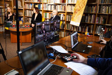 Grupa osób w bibliotece uczestniczy w wydarzeniu online, przed laptopami zamieszczonymi na stołach, z mikserem audio na pierwszym planie. Osoba w tle przemawia do mikrofonu, obok niej stoją banery i eksponaty kolejowe. W tle widoczne są półki z książkami i zdjęcie pociągu na ścianie.