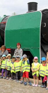 Grupa dzieci w jaskrawych kamizelkach odblaskowych stoi w linii obok dużego zielonego parowozu z czerwonymi elementami. Obok dzieci znajdują się trzy osoby dorosłe, również pozujące do zdjęcia. W tle są zielone krzewy i niebo pokryte chmurami.