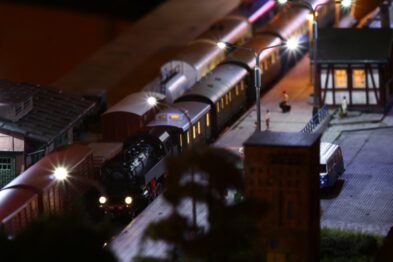 Pokazano model stacji kolejowej z pociągiem i peronami oświetlonymi latarniami, tworzącymi realistyczne światło. Detale takie jak wagony, budynki i ludzkie postacie są starannie odwzorowane. Makietę cechuje nocna atmosfera, co podkreślają cienie i punktowe źródła światła.