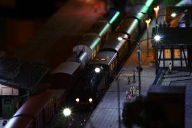 Zminiaturyzowana lokomotywa parowa z wagonami stoi na torach obok peronu, oświetlona przez punktowe lampy. Postacie w skali modelu znajdują się na peronie i wzdłuż torów, w roli pasażerów i pracowników kolei. Scena jest stylizowana na nocną, zaś na torach i makiecie widać szczegóły takie jak budki, ławki i oznakowania kolejowe.